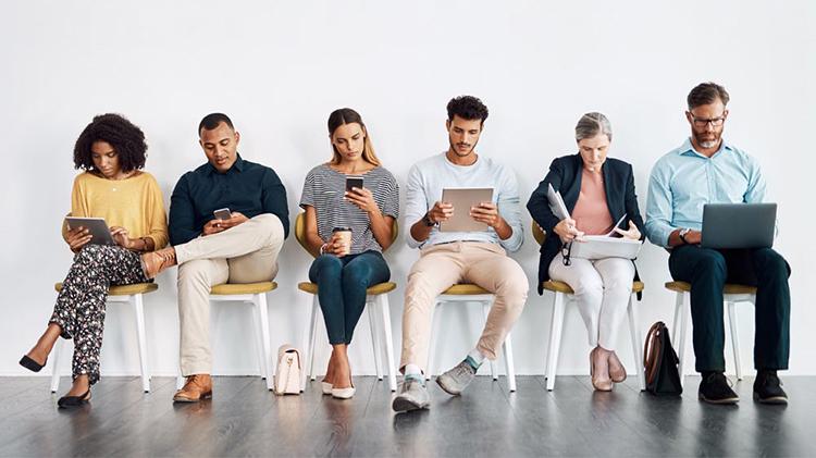 Six people sitting in a row of chairs looking at their laptops, smartphones and tablets.